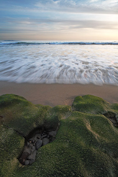 The incoming rush of the ocean seems to be reaching out to this green rock formation.