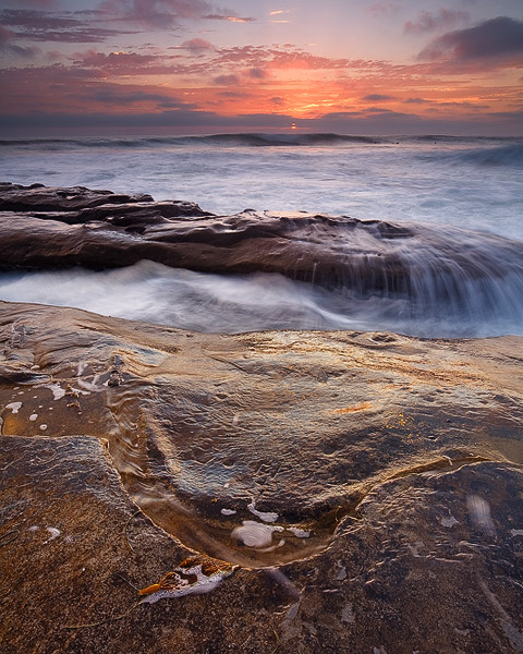 High tide washes over shoreline rocks, as the sun slides in between the horizon and some low clouds.