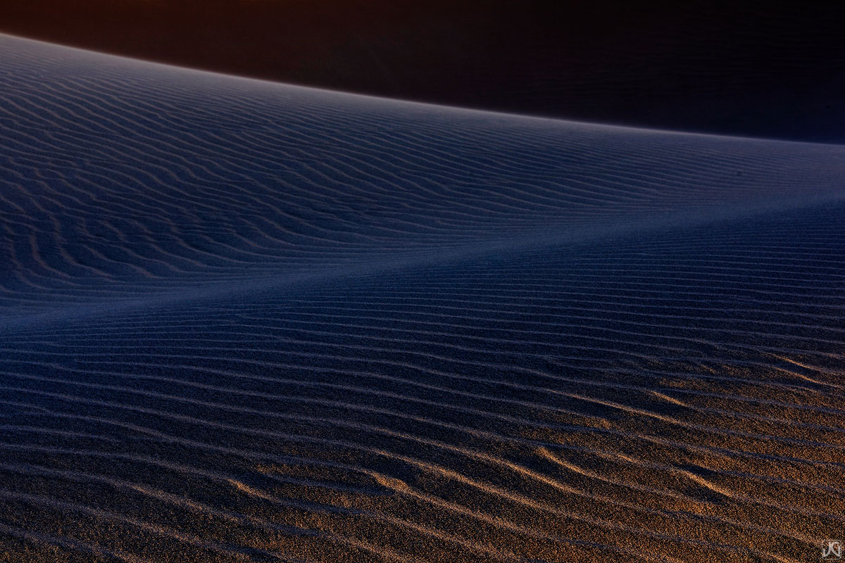 Late afternoon light creeps over the dunes, illuminating the ridges and patterns in the sand, while casting cool toned shadows...