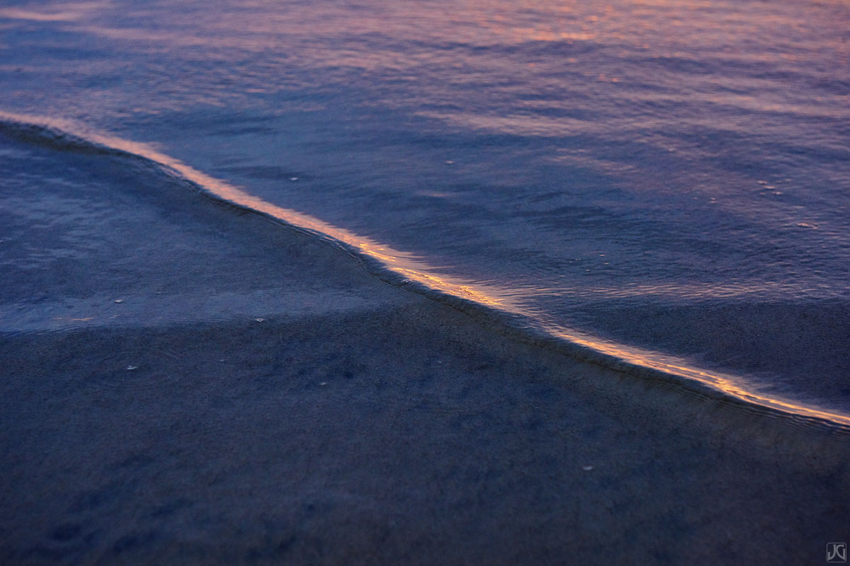 A small wave travels slowly towards the beach in the shallow waters along the coast at sunset.