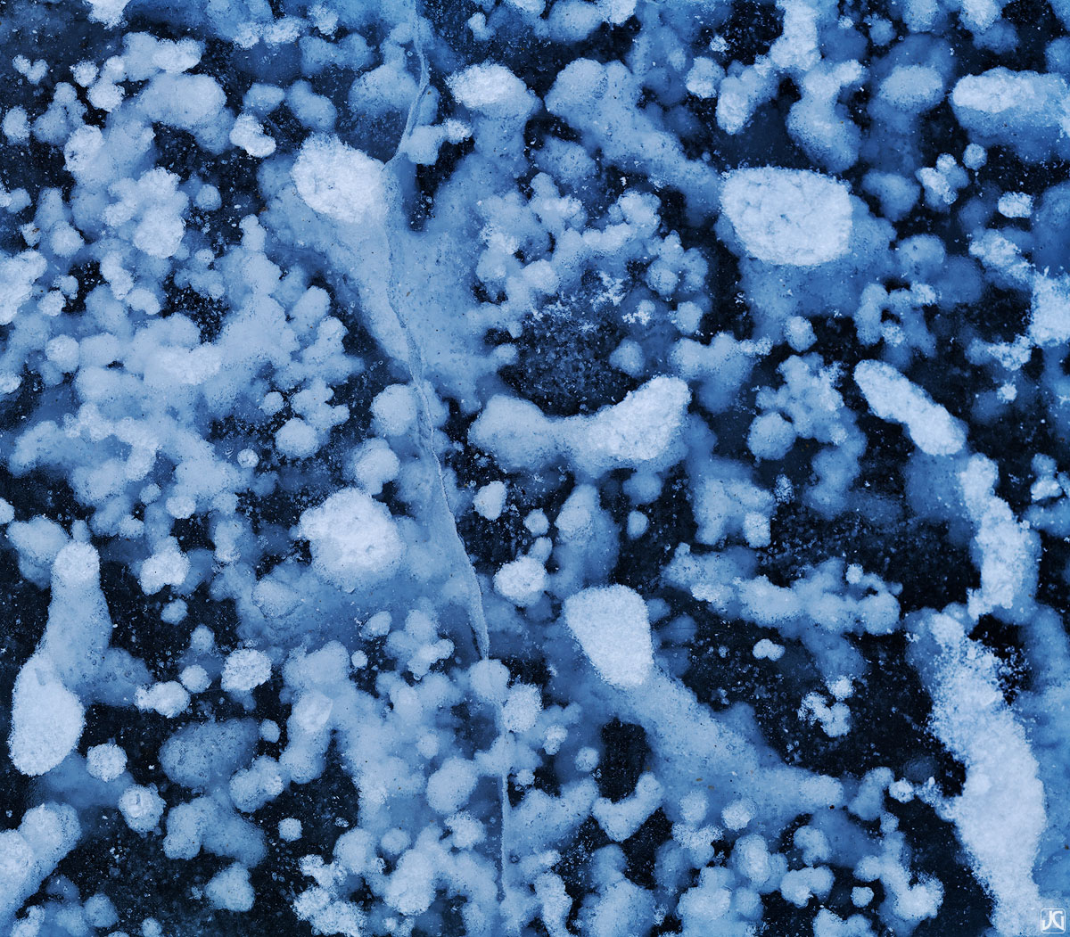 Snow, bubbles and cracks form a unique network of shapes and patterns throughout the ice of a frozen lake.